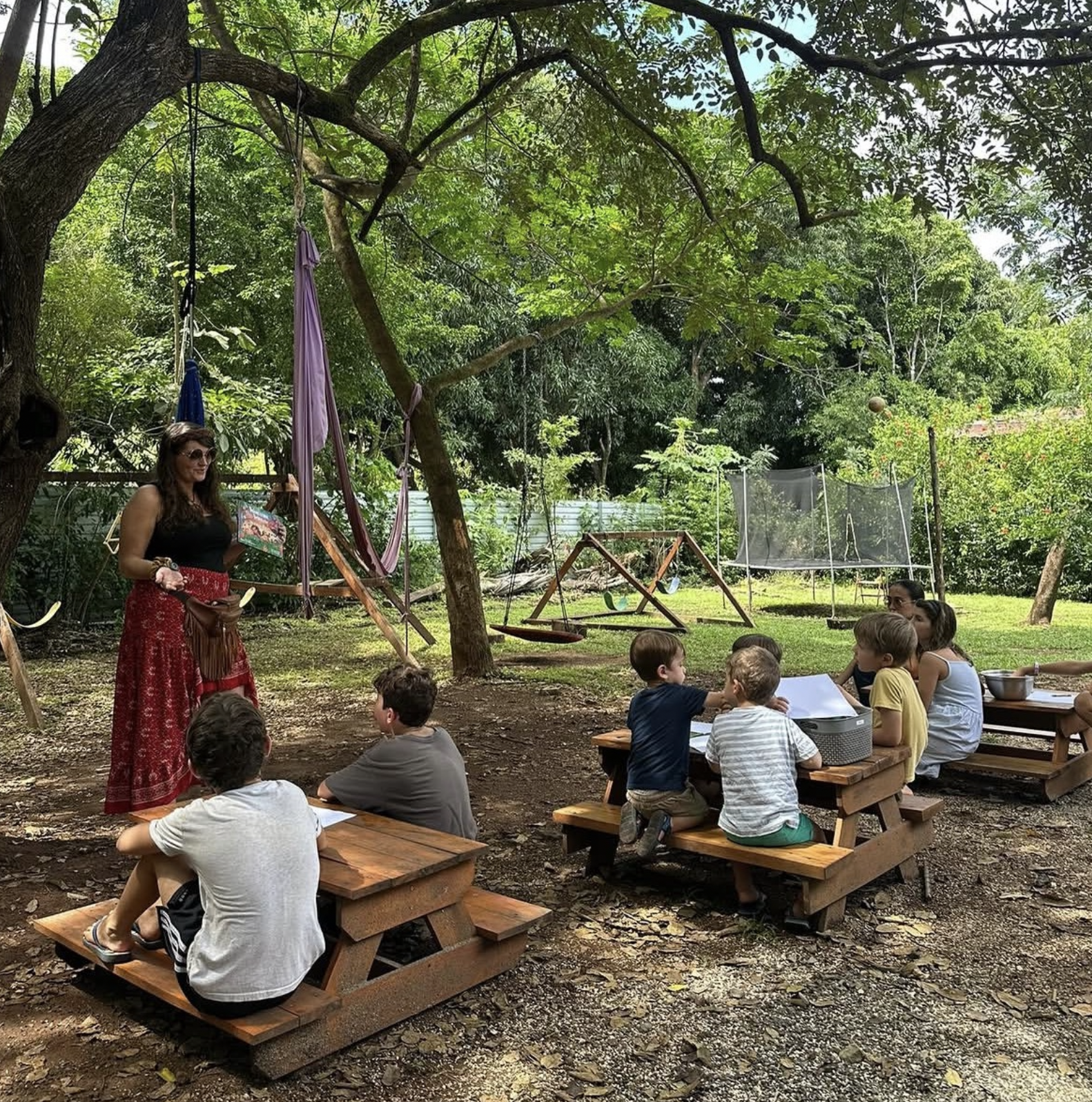 Children and parents gathered at outdoor picnic-table desks for a lesson under the trees at a local education setting
