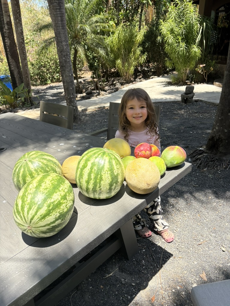 A young girl beaming beside a table piled high with fresh watermelons, melons and mangoes