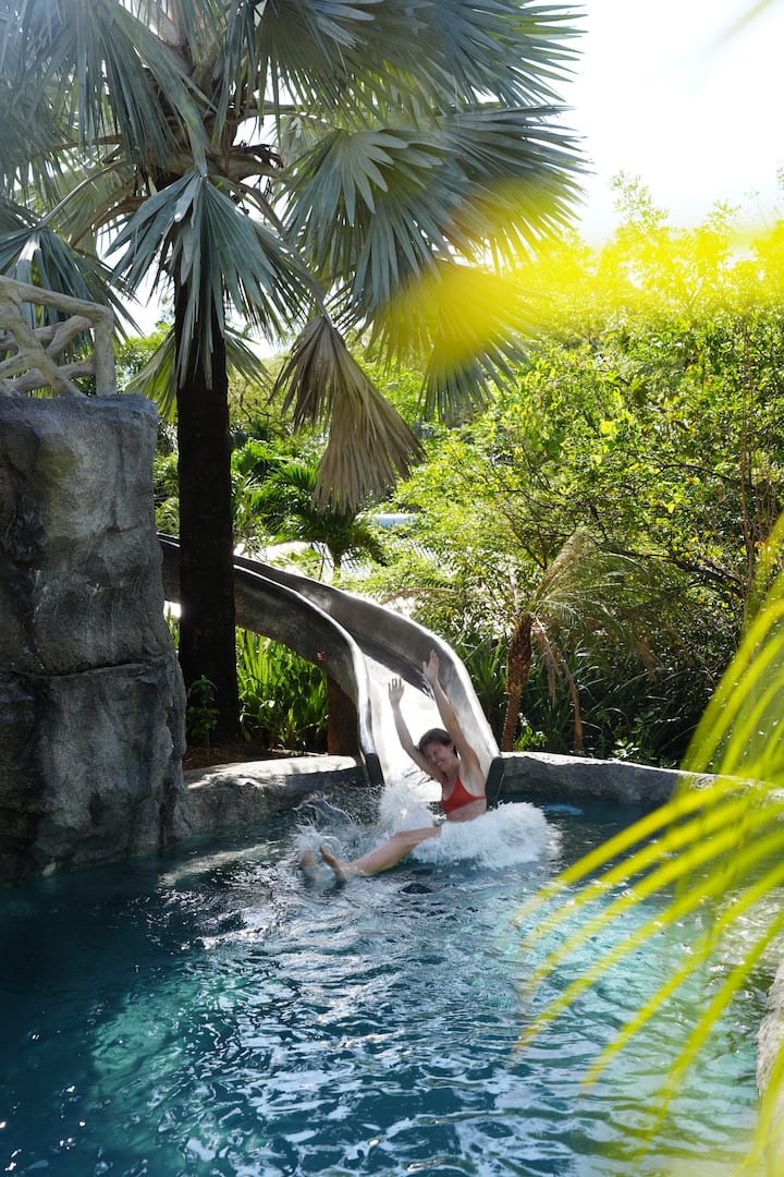 A child splashing down the water slide into the hotel pool, framed by sunlit palms