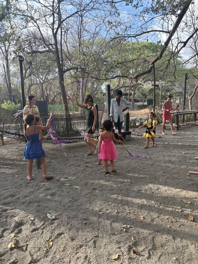 A group of children, one in a pink dress, dancing with ribbon wands together under the trees